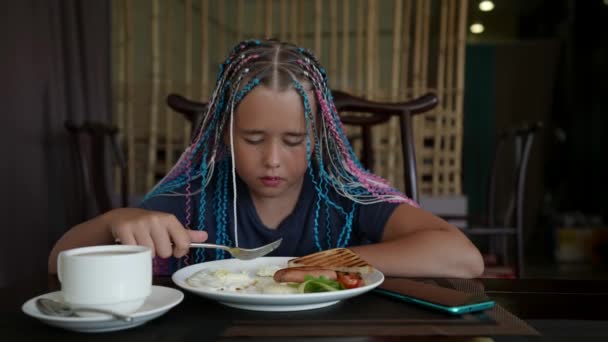 Un adolescent prend son petit déjeuner et regarde une vidéo sur son téléphone mangeant des œufs frits avec une saucisse — Video