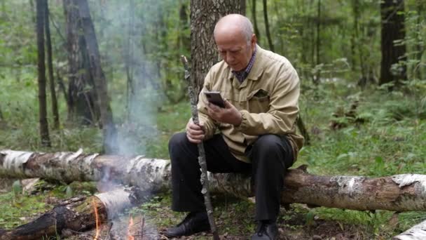 Ein Rentner sitzt im Wald am Feuer auf einem Baumstamm und kommuniziert per Telefon mit der Außenwelt — Stockvideo