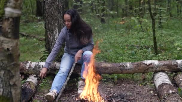 Жінка-туристка гріється біля багаття в лісі, щоб кинути гілку, щоб підтримувати полум'я — стокове відео