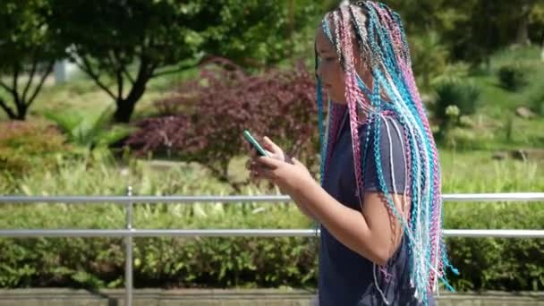 Beyaz bir genç kız cep telefonu kullanıyor, yaz parkında yürüyor. Açık havada akıllı bir telefonla internette sörf yapan sevimli bir gencin portresi. — Stok video