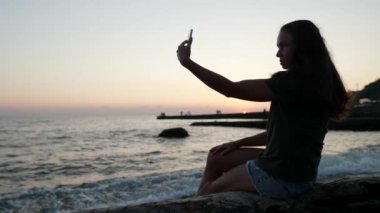 Plaj siluetinde selfie çekerken mutlu ol. Akıllı telefonlu ince bir kadın deniz sahilinde gün batımında cep telefonunun fotoğraflarını çekiyor.