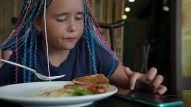 Saçları örgülü şık bir genç kız bir restoranda bir masada oturmuş kahvaltı yapıyor ve telefonundan sosyal ağları izliyor.
