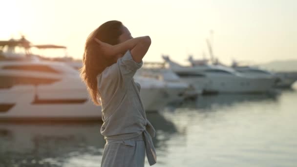 Een mooie natuurlijke vrouw staat bij de zeehaven bij zonsondergang in de avond recht haar haar in de stralen van de zon — Stockvideo