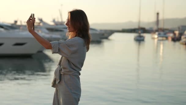 Nette natürliche Frau, die abends bei Sonnenuntergang Fotos mit ihrem Handy macht, steht am Hafen — Stockvideo