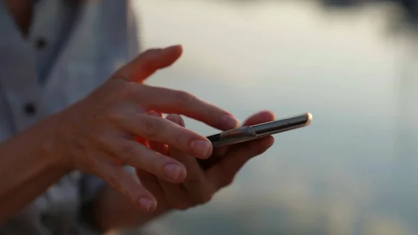 Les mains de la femme tiennent le téléphone et déplacent son doigt sur l'écran tactile dans la lumière du coucher du soleil dans la rue Image En Vente