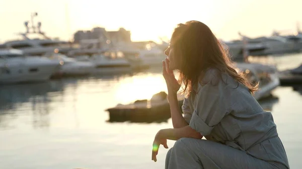 Portrait d'une femme assise sur la plage avec des yachts sur le fond dans les rayons du soleil couchant et triste Images De Stock Libres De Droits