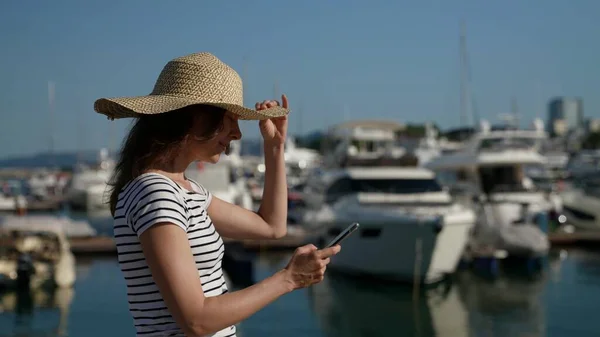 Hezká dáma s velkým slamákem kráčející podél moře s jachtami používá svůj telefon k zobrazení sociálních sítí Stock Obrázky