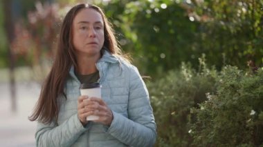 Yorgun, düşünceli, orta yaşlı bir kadın güneşli bir sonbahar gününde elinde bir fincan sıcak kahveyle sokakta oturuyor.