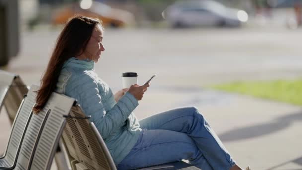Μια χαρούμενη ενήλικη γυναίκα με ένα τηλέφωνο στα χέρια της στο δρόμο το φθινόπωρο να πίνει καφέ. — Αρχείο Βίντεο