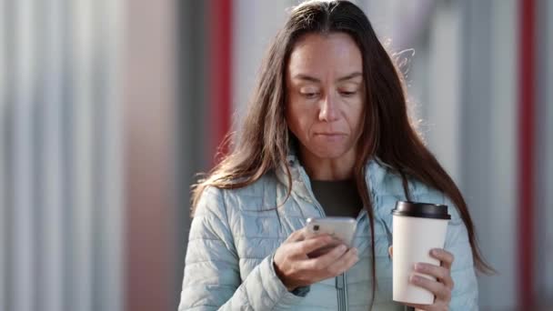 Ritratto di una donna entusiasta sorpresa in piedi per strada con un telefono che sfoglia un feed di social network e tiene in mano una tazza di carta con caffè — Video Stock
