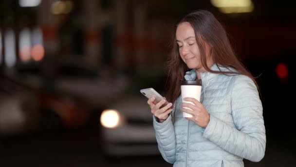 Retrato de una mujer alegre de mediana edad en un estacionamiento con un teléfono en la mano hojeando una fuente de noticias y bebiendo café de una taza de papel — Vídeos de Stock