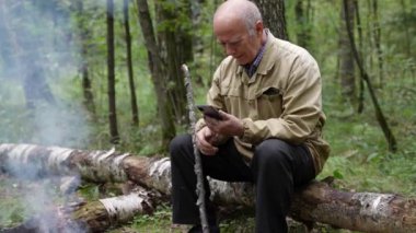 Ormanda ateşin yanında oturan yaşlı bir adam bir kütüğe oturup internete erişmek için telefonunu kullanıyor.