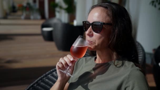 Una feliz dama de éxito en gafas de sol está sentada en la terraza de un restaurante y bebiendo una copa de champán rosa frío — Vídeos de Stock