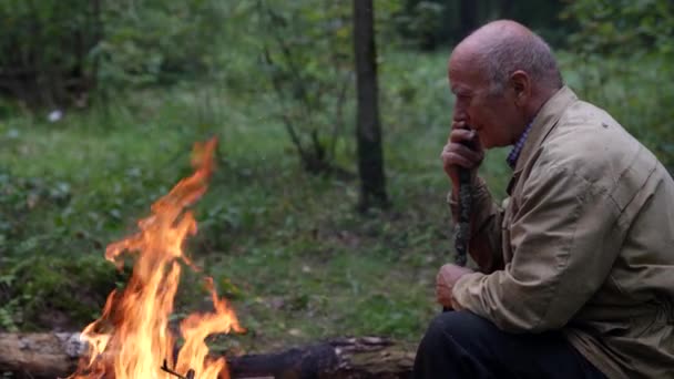 Un vieil homme réfléchi est assis dans la forêt en face du feu et se souvient de la vie qu'il a vécue, le regard sage du vieil homme — Video