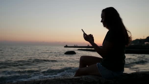 Silhouette une femme est assise sur l'océan avec un téléphone qui écrit un message texte et passe la journée au resort — Video