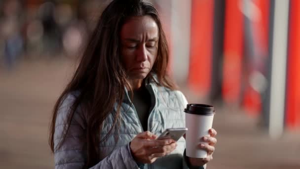 Uma mulher adulta atraente e elegante Usando seu smartphone, segurando uma xícara de café de papel, de pé contra o fundo de uma cidade moderna. — Vídeo de Stock