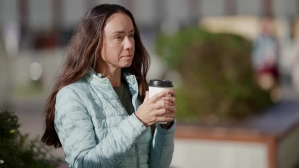 Привлекательная взрослая женщина пьет кофе из бумажной чашки, сидя на скамейке на городской улице в осеннем парке — стоковое видео