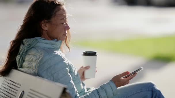 Porträtt av en kvinna med mobiltelefon och pappersmugg. En vuxen kvinna skriver SMS på en smartphone och dricker hämtkaffe. Attraktiv kvinna med mobiltelefon och varm dryck utomhus — Stockvideo