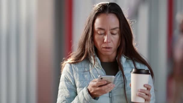 Una donna adulta beve caffè da una tazza di carta in una strada della città guarda i messaggi di testo su uno schermo del telefono cellulare — Video Stock