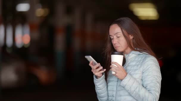 Μια γυναίκα κρατάει ένα χάρτινο φλιτζάνι καφέ στο πάρκινγκ. Καφές για να τον πάρεις. Μια κομψή ενήλικη γυναίκα δακτυλογραφεί μηνύματα στο smartphone της και πίνει καφέ από ένα χάρτινο κύπελλο — Αρχείο Βίντεο