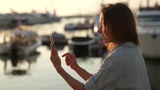 Çekici, orta yaşlı bir kadın kumsalda, batan güneşin altında elinde bir telefonla oturmuş test mesajları yazıyor. — Stok video