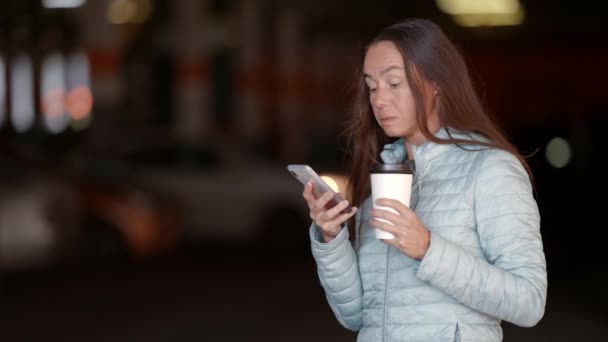Atractiva mujer adulta tiene café, utiliza el teléfono mientras está de pie en un aparcamiento — Vídeos de Stock
