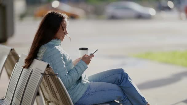 Cep telefonu ve kağıt bardağı olan bir kadının portresi. Genç bir kadın akıllı telefonuyla mesaj atıyor ve paket kahve içiyor. Cep telefonu ve açık havada sıcak içeceği olan yetişkin bir kadın. — Stok video