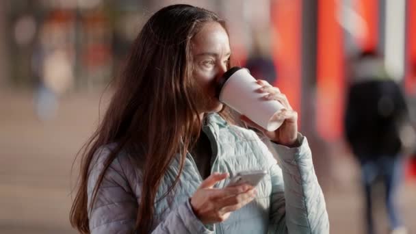 コーヒーを片手に路上に立つジャケット姿の女性が電話でメッセージを書いている。都市生活の概念 — ストック動画