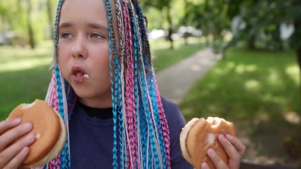 Une adolescente élégante mange avec gourmandise deux hamburgers de restauration rapide. manger dehors pendant la journée en été. — Video