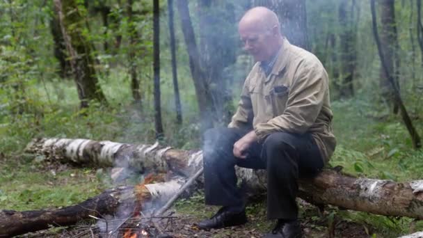 Um homem velho na floresta está sentado em um tronco perto do fogo um monte de fumaça está voando em sua cara — Vídeo de Stock