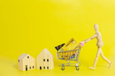 Ahşap manken ya da kukla alışveriş arabası, kredi kartı ve ahşap ev, sarı arka plan, iş ve mülk konsepti..