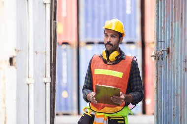 Foreman üniforma giyiyor ve konteynır kargosunda pano ile lojistik kontrolleri yapıyor, konteynırların yükleme alanı lojistik ihracat ve nakliye işlemlerini kontrol ediyor..