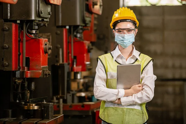 woman engineering wear protection face mask and safety helmet holding tablet standing front machine in factory Industrial. new normal work during epidemic covid-19.
