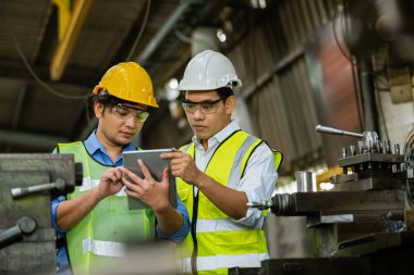 Two asian engineering man wearing safety uniform and hard hats working with laplet work machine lathe metal, Heavy industry worker man concept.