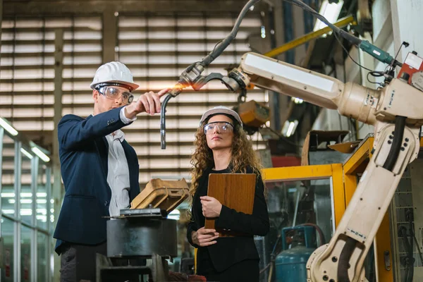 man engineer and female assistant wearing black suit and white hard hat in factory male engineering using automatic box controls robotic arms machine. industrial technology concept.