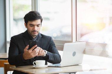 İş adamı, kafeteryada otururken akıllı telefon kullanarak siyah takım elbise giyer. Yakışıklı erkek işçi dinlenme zamanı..