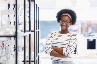 Afro-Amerikan saçlı genç kadın portresi gülümsemesi ve çapraz eller optik dükkanında duran gözlük takıyor. modern göz doktoru konsepti.