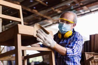 Yaşlı marangoz koruyucu maske takıyor ve kulaklık takıyor. Odun cilalamak için zımpara kağıdı kullanıyor. Fabrika endüstrisi ahşap işi.