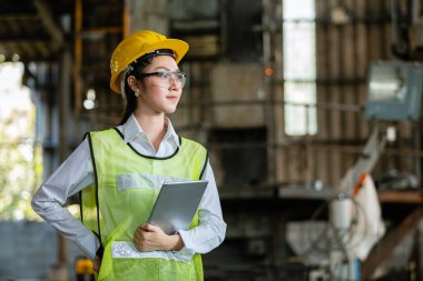 Asyalı kadın mühendis sanayii ağır işçisi, baret takıyor ve tablet tutuyor fabrikada makine alanında duruyor, mühendislik endüstrisi kavramı.