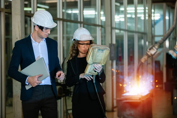 man engineer and female assistant wearing black suit and white hard hat in factory male engineering using automatic box controls robotic arms machine. industrial technology concept.