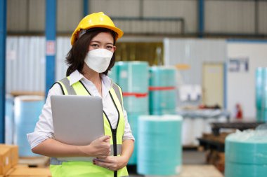 Kadın mühendis koruyucu yüz maskesi takıyor ve depo fabrikasında dizüstü bilgisayar tutuyor. Hijyen maskesi endüstrisi üretimi.