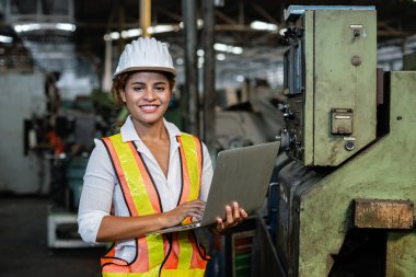 Üniforma giyen Afrika kökenli kadın işçiler güvenlik ve başlık takarak sistem makinelerini fabrikada dizüstü bilgisayarla kontrol ediyorlar. Büyük sanayi fabrikasındaki kadın..