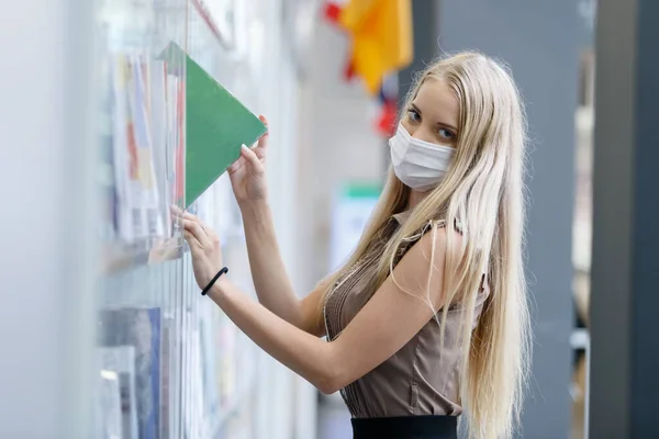 金髪の髪の美しい女性は 図書館で医療マスク立って本棚を身に着けています 教育の概念 — ストック写真