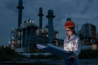 Elektrik santralinde gece vardiyası sırasında güvenlik kaskı takan kadın sanayi mühendisi, mühendislik ve inşaat sektöründe liderlik, planlama ve çeşitliliği sembolize ediyor..