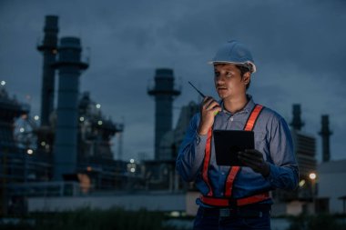 Petrol rafinerisi veya elektrik santralinde gece vardiyası teftişi sırasında telsiz ve dijital tablet kullanarak güvenlik üniforması giyen endüstri mühendisi, sistem güvenliği ve işlemleri sağlıyordu..