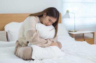 Asyalı bir kadın yatakta yastığa üzgün bir ifadeyle sarılıyor, depresyon konsepti, yalnızlık, duygusal hassasiyet ve sakin ve sessiz bir yatak odasında zihinsel sağlık farkındalığı..