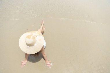 Hasır şapkalı, kumlu tropikal plajda dinlenen bir kadın, yukarıdan bakıldığında, huzurlu ve asgari yaz manzarası konsepti. Seyahat, tatil, rahatlama ve kıyı yaşam tarzı görselleri için ideal.