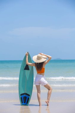 Kadınlar yaz tatilinin tadını sörf tahtasıyla çıkarıyorlar. Turuncu hasır bluz ve hasır şapka giyiyorlar. Güneşli tropikal ortamda gülümsüyorlar. Yaşam tarzı, seyahat ve yaz tatili temaları için mükemmel..
