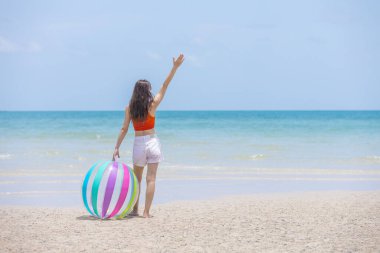 Genç bir kadın kumsalda renkli bir plaj topuyla okyanusa bakıyor ve denize doğru el sallıyor. Yaz tatili, özgürlük ve tropikal plaj yaşam tarzı kavramları için mükemmel..