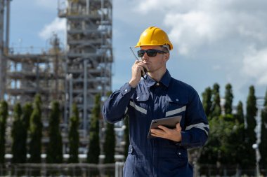 Endüstriyel santral bölgesinde telsiz ve tablet kullanarak güvenlik kaskı takan mühendis, iletişim, güvenlik yönetimi, teftiş süreci ve rafineri petrol fabrikası işletmesini temsil ediyor..
